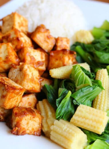 closeup image of tofu coated in peanut sauce with a side of rice and sauteed bok choy with chopped baby corn