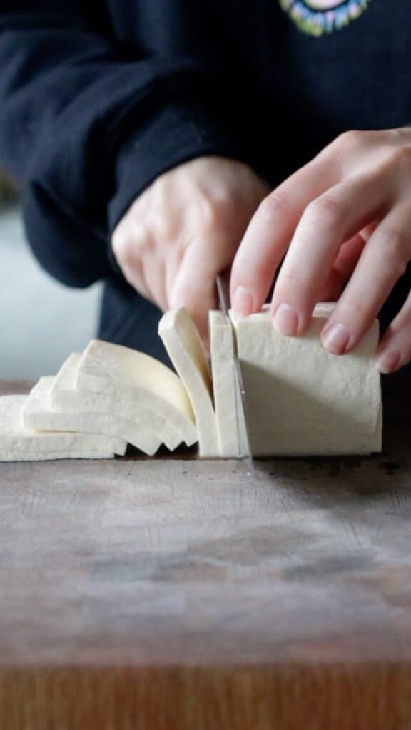 half of a block of tofu being thinly sliced on a wooden cutting board