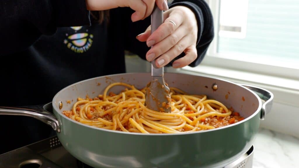 bucatini pasta being mixed in with the tofu bolognese sauce in a sauté pan