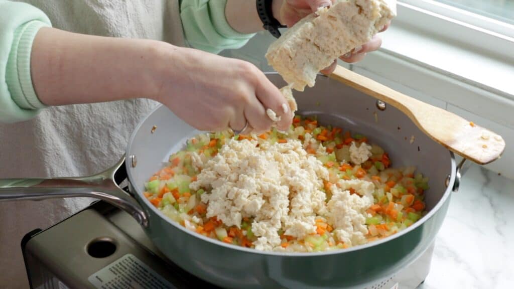 tofu being crumbled into a pan full of sautéing diced carrot, onion, and celery
