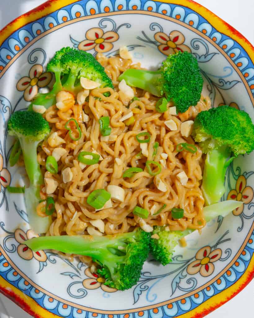 Close-up overhead shot of Peanut Butter Noodles in a bowl. There are florets of broccoli around the outside of the bowl and the noodles are topped with chopped peanuts and scallions.