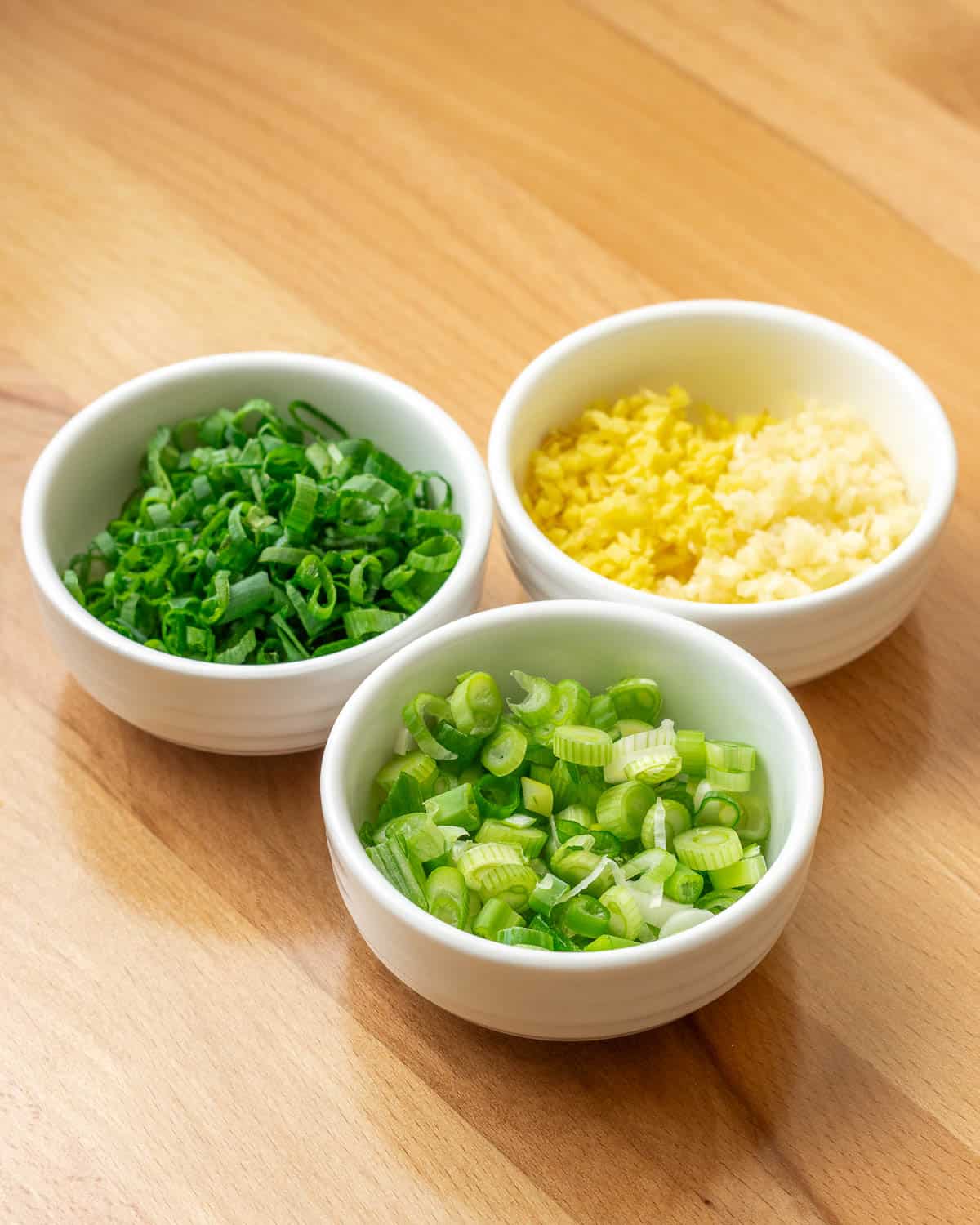 three white pinch bowls containing scallions, garlic, and ginger