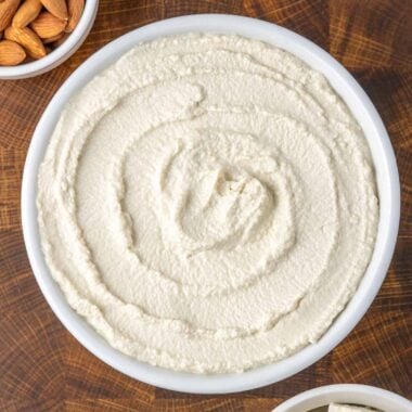overhead shot of white bowl of vegan ricotta next to a bowl of cubed tofu, a small bowl of almonds, and a white kitchen towel