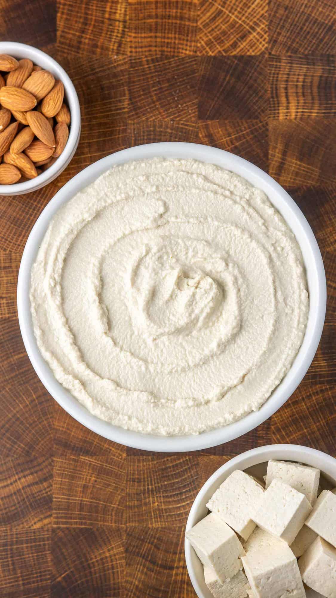 overhead shot of white bowl of vegan ricotta next to a bowl of cubed tofu, a small bowl of almonds, and a white kitchen towel