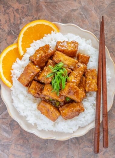 a plate of vegan orange tofu served over white rice, with chopsticks and two orange slices