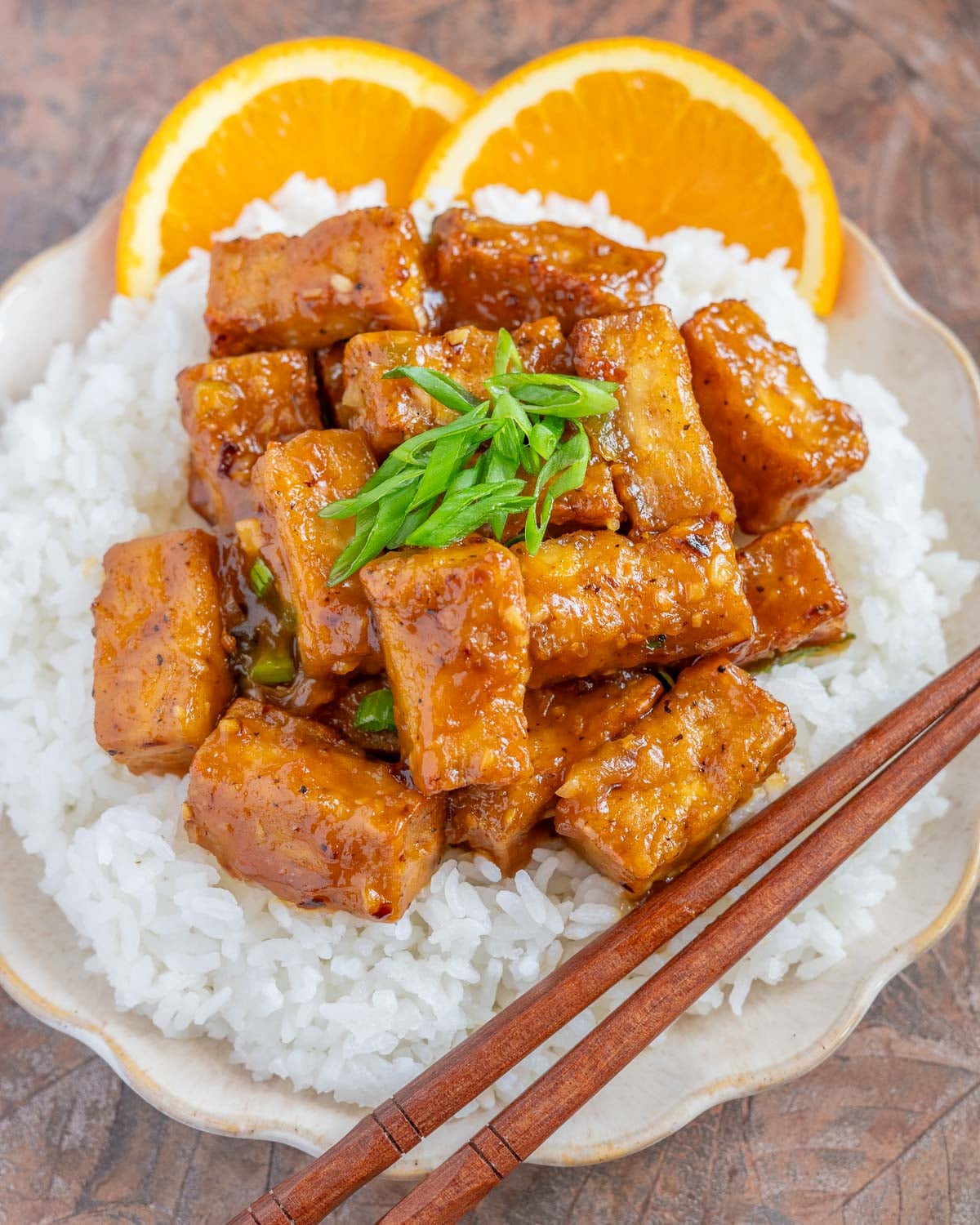 a plate of vegan orange tofu served over white rice, with chopsticks and two orange slices