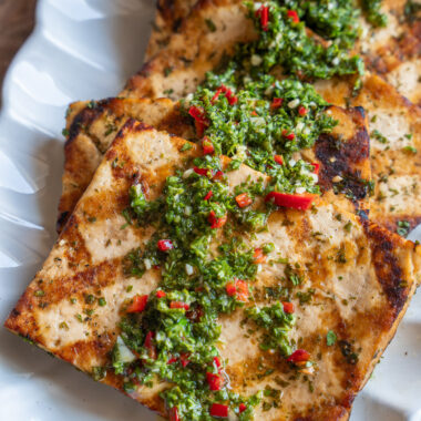 steaks of grilled tofu lined up on a plate and topped with chimichurri