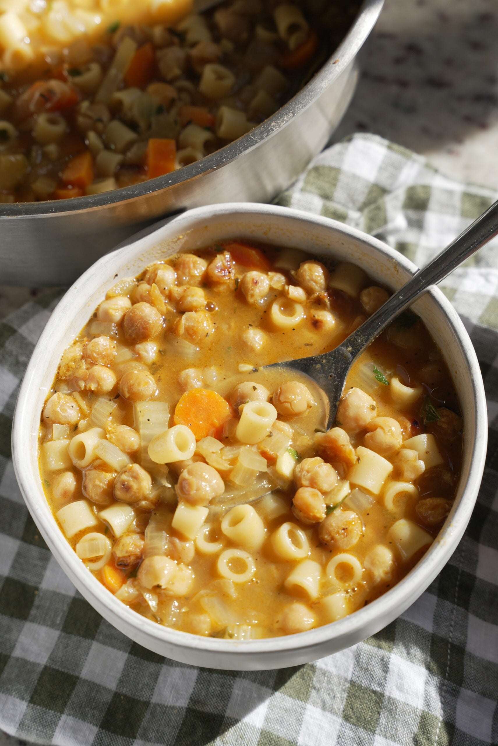 Chickpea noodle soup in a bowl on a green and white checkered towel. In the background is the full pot of soup with a ladle.